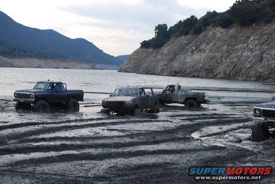Four-wheel drive trucks stuck in mud near a lake, one blue pickup in foreground, others further back