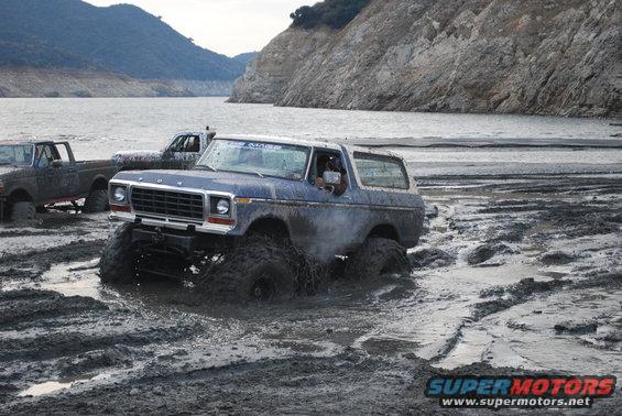 Blue Ford Bronco with huge tires stuck in mud, another truck behind it