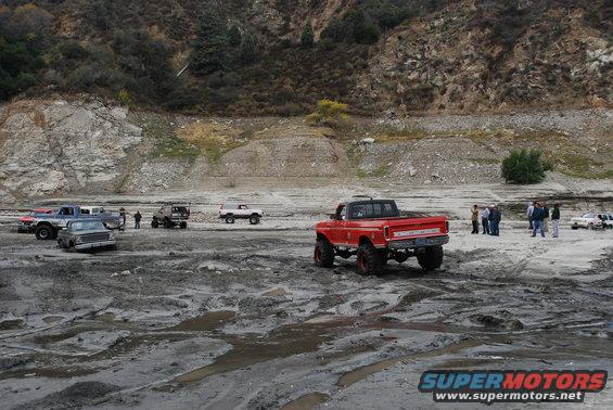 Red pickup truck with lifted suspension and large tires in a muddy area. Other trucks and SUVs are visible in the background