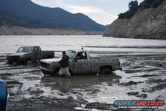 Two trucks stuck in mud with a person pushing one. Water and hills in the background