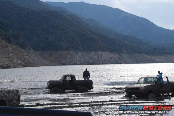 Two trucks are stuck in the mud by a lake with mountains in the background. One truck has a person standing in the bed