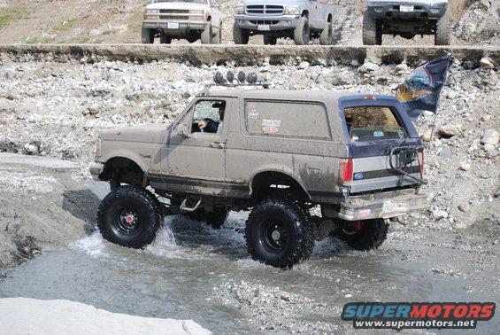 Muddy tan and blue Ford Bronco with lifted suspension and large tires driving through water
