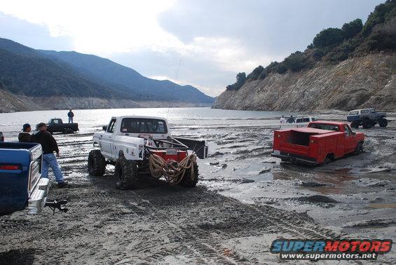 Several trucks, including a white pickup with large tires and a red utility truck, stuck in mud near a body of water with hills in the background