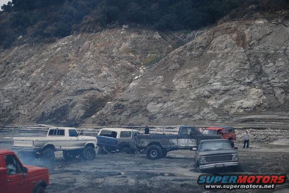 Several trucks and SUVs are parked on a muddy, rocky terrain with a large, barren hill in the background