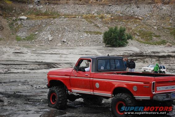 Red lifted pickup truck with black top, side view. Other vehicles in background