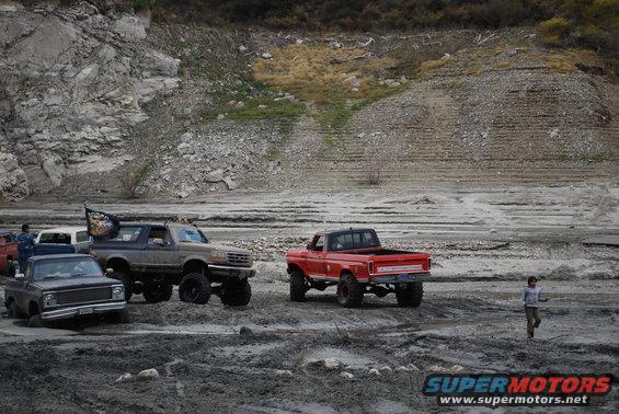 1996 Ford Bronco with flag, red pickup truck, and dark pickup truck in mud