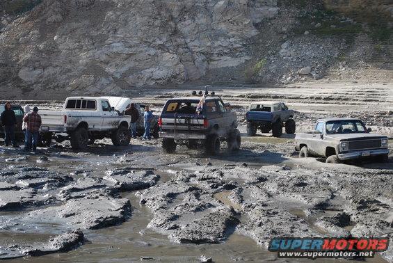 Several lifted trucks and SUVs stuck in thick mud near a rocky hillside