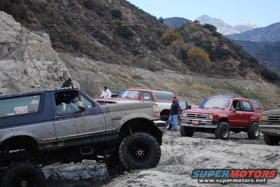 Mud-splattered blue and grey Ford Bronco with lifted suspension and large tires, parked on a rocky, off-road terrain