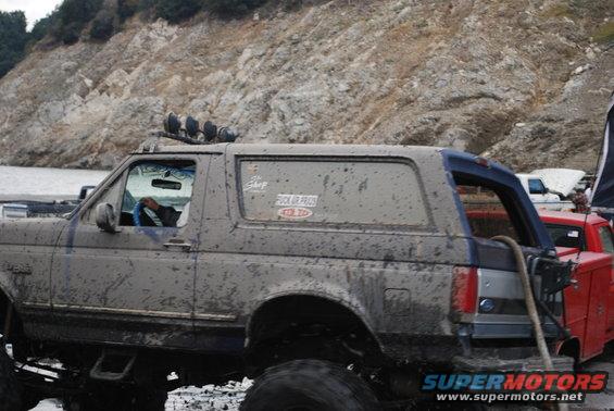 Muddy SUV with off-road lights on roof, driving in a rocky area. Another red truck is visible