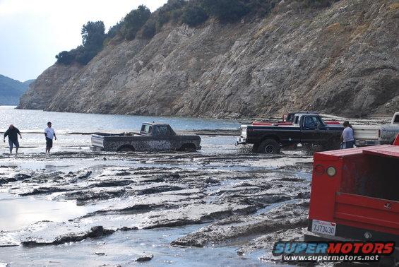 Several trucks stuck in mud near a body of water with a rocky hillside