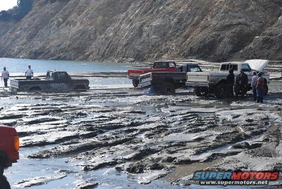 Several trucks stuck in deep mud near a body of water and a rocky hill