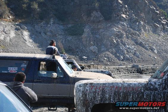 Mud-covered dark blue and tan Ford Bronco with a person sitting on the roof