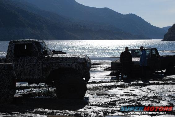 Two mud-covered trucks parked on a muddy shore next to a body of water with mountains in the background