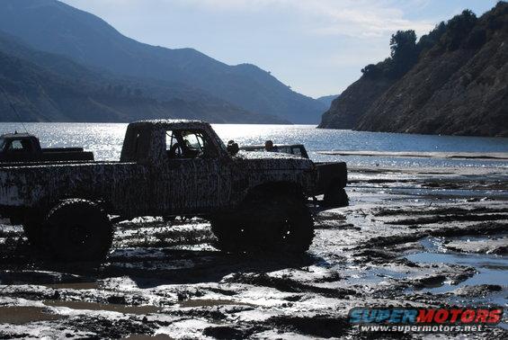 Mud-covered pickup truck stuck in mud near a body of water, with mountains in the background