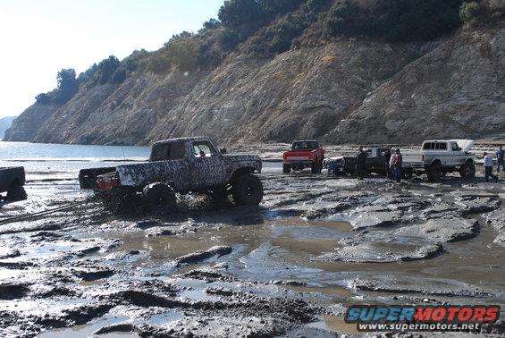 Several trucks stuck in mud near a body of water with a rocky hillside in the background