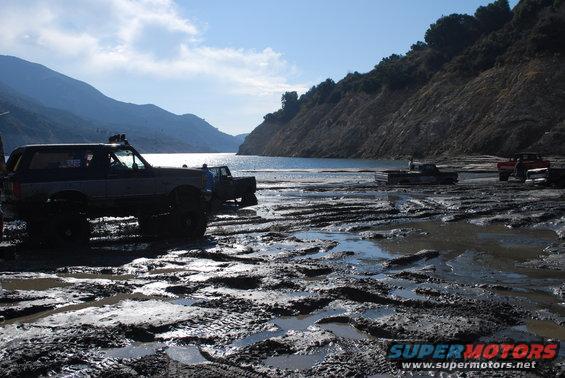 Dark blue Ford Bronco with off-road tires parked on muddy ground near a body of water
