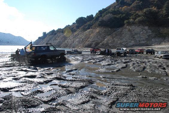 Multiple off-road vehicles navigating a muddy, wet terrain with a large body of water and hills in the background