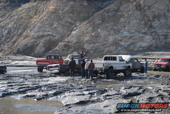 Several trucks and a Bronco stuck in thick mud with people around