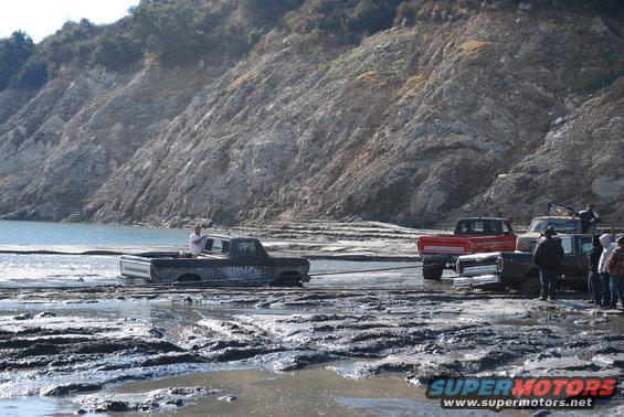 Muddy scene with several trucks stuck in thick mud near a body of water and a rocky hill