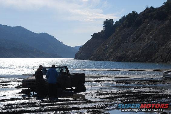 Muddy truck stuck in mud near water with two people standing beside it