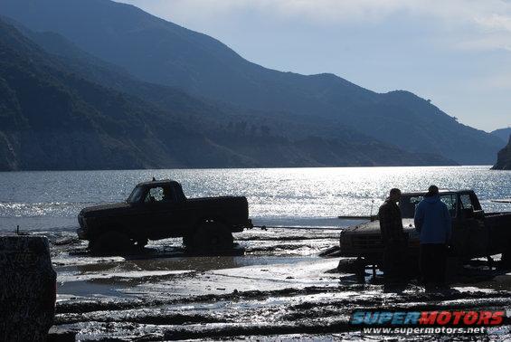 Two pickup trucks stuck in mud near a body of water with mountains in the background
