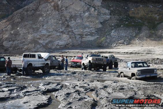 Several lifted trucks stuck in thick mud near a rocky hillside