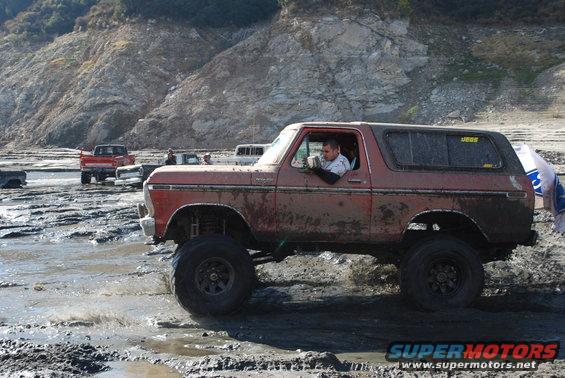 Mud-covered 1978 Ford Bronco with lifted suspension driving through a muddy terrain. Another red truck is visible in the background