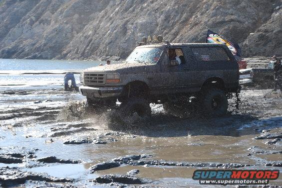 Muddy lifted SUV splashing through mud, 3q-front view. Visible text 'SUPERMOTORS'