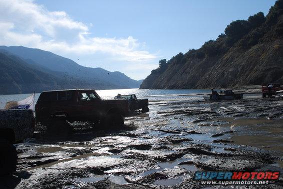 Muddy off-road vehicles driving through a wet, muddy area near a body of water and hills