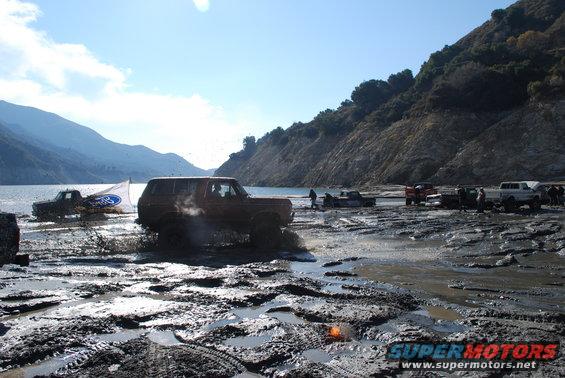 Brown Ford Bronco driving through mud with a Ford flag on another truck in the background