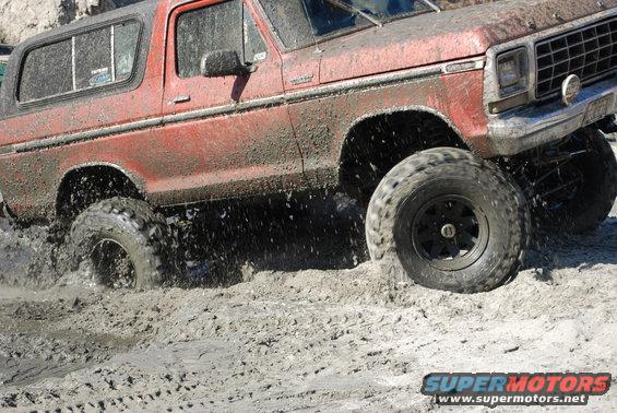 Red SUV with large tires splashing through mud. Visible text: SUPERMOTORS.NET