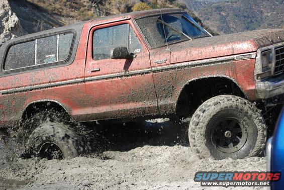Red Ford Bronco with lifted suspension driving through thick mud, splashing water
