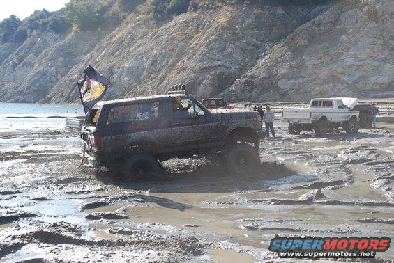 Mud-splattered dark blue Ford Bronco with a flag on the antenna, stuck in mud with front wheels turned