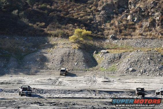 Three trucks on a dry, dusty landscape with hills in the background. One truck is in the foreground, another in the mid-ground, and a third further back