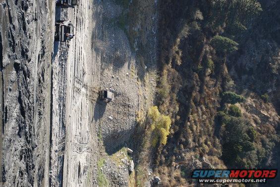 Several off-road vehicles driving on a muddy, rutted track next to a steep, vegetated hillside