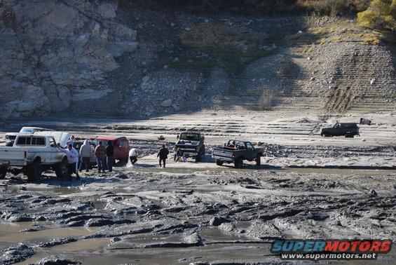 Several trucks and SUVs are stuck in a muddy, dry lake bed with people around them