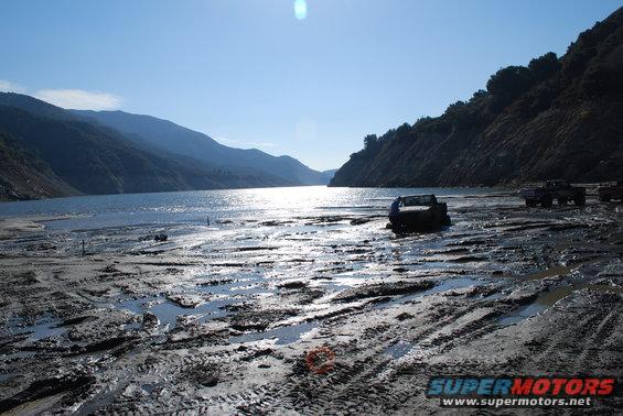 Several trucks stuck in mud near a lake with mountains in the background