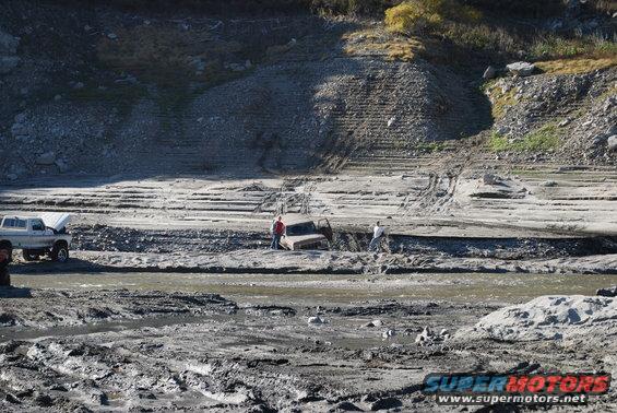 Two vehicles stuck in mud, one with hood open. People are standing near them