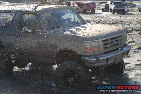 1996 Ford Bronco covered in mud, driving through water with other vehicles in the background