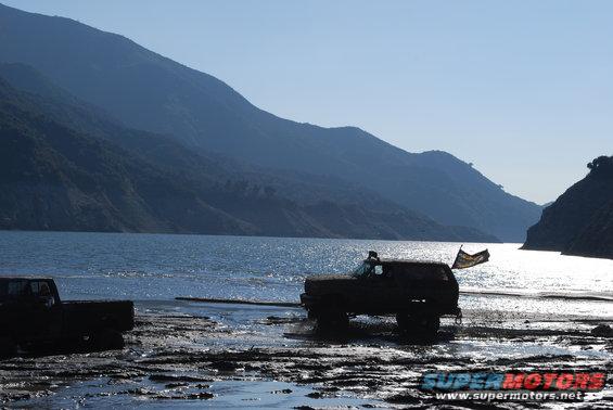 Dark 1992 Ford Bronco with a flag on the back, partially submerged in muddy water near a lake