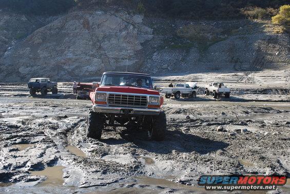 Red pickup truck with lifted suspension and large tires in mud, front view. Other trucks visible in background