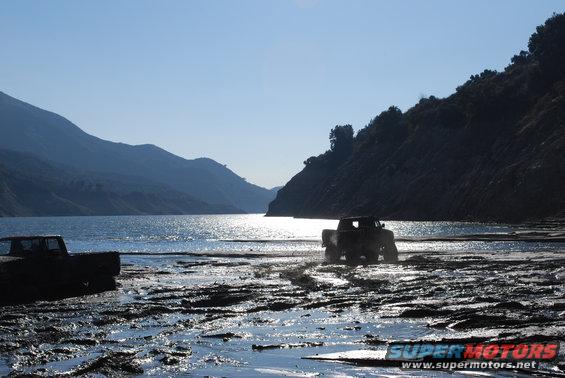 Two trucks are in a muddy, watery area near a lake with mountains in the background