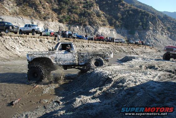 Muddy pickup truck driving through water and dirt. Other trucks and people are on a ridge in the background