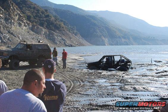 Two off-road vehicles stuck in mud near a lake with mountains in the background