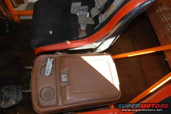 Brown plastic center console with cup holders and a latch, next to a black seat with red trim and a roll cage