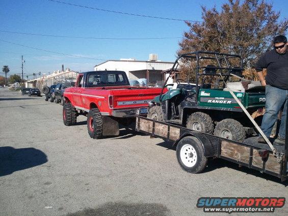 Red pickup truck with large tires towing a trailer carrying a green Polaris Ranger UTV
