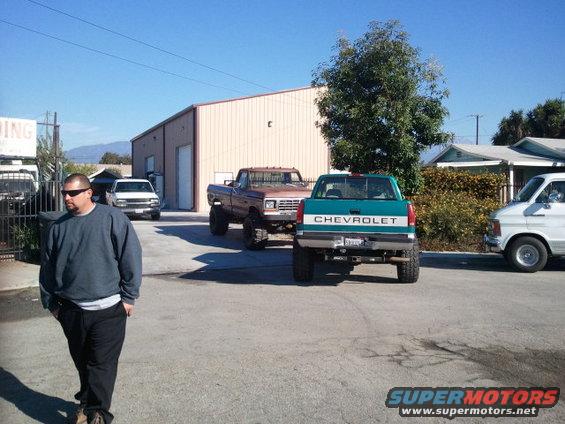 Teal Chevrolet pickup truck, lifted, rear view. Brown Ford pickup truck behind it. White van to the right