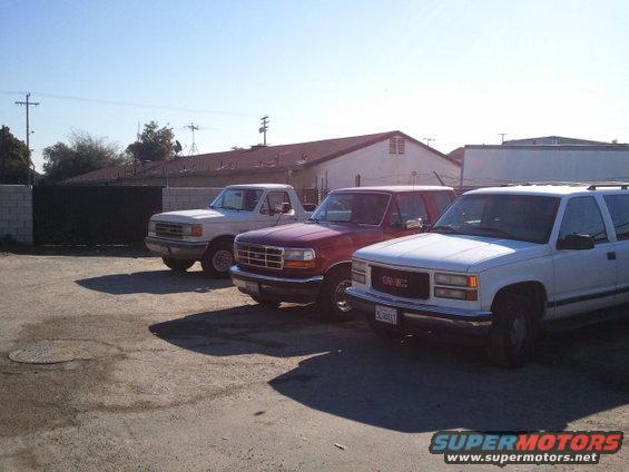 Three trucks parked in a lot: a white Ford F-Series, a red Ford Bronco, and a white GMC Suburban