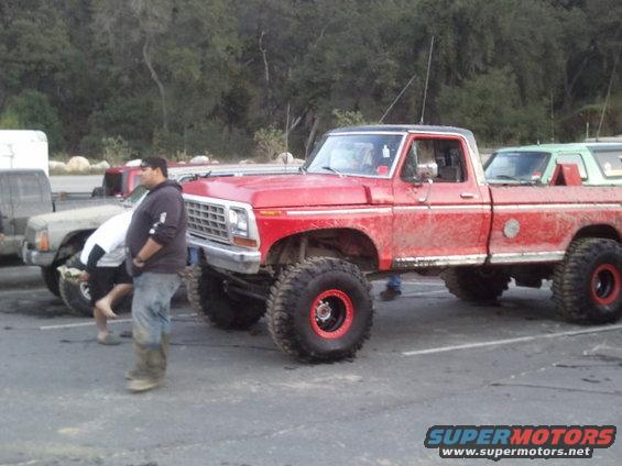 Red lifted pickup truck with big tires parked next to another truck. Someone is looking under the hood of the other truck