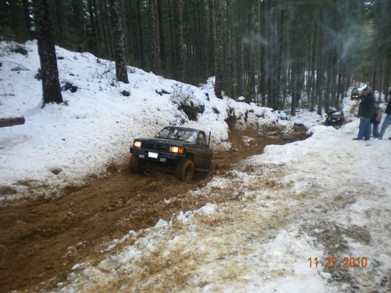 Pickup truck driving through a muddy trail with snow on the sides. Other vehicles and people are visible further down the path in a wooded area